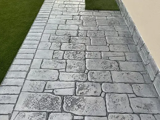 Schmaler Hauszugang aus grauem Prägebeton entlang einer Rasenfläche