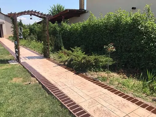 Gartenweg aus Prägebeton mit Ziegelkante, der unter einem Rosenbogen hindurchführt