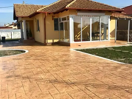 Grosse Hoffläche aus sandfarbenem Prägebeton vor einem Bungalow mit Veranda